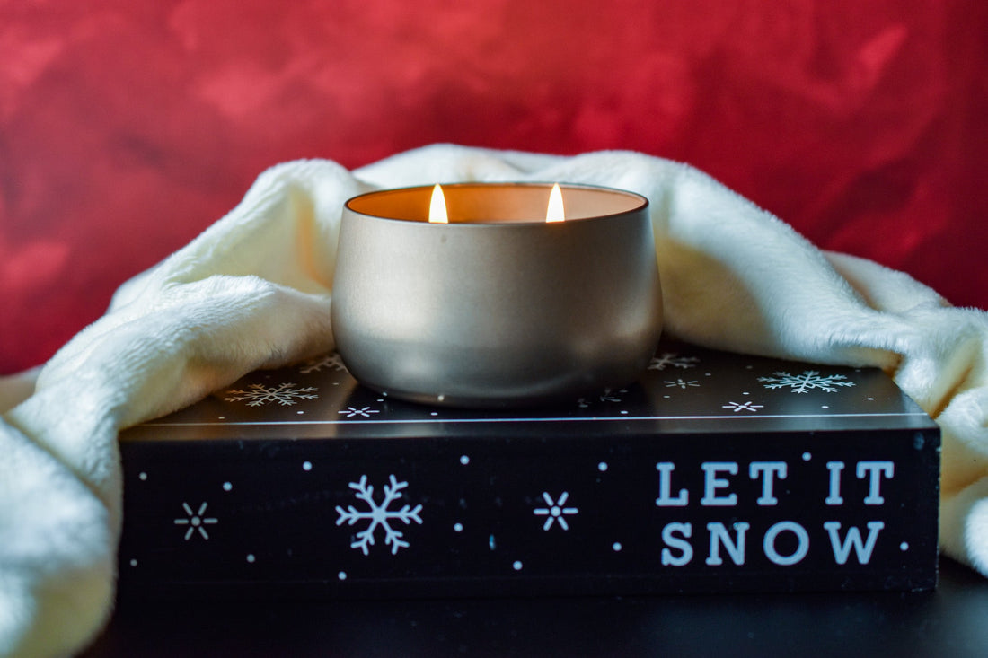 Christmas in July candle. Lit Gold bubble candle tin sitting on a prop that says Let it Snow. Red bakcground