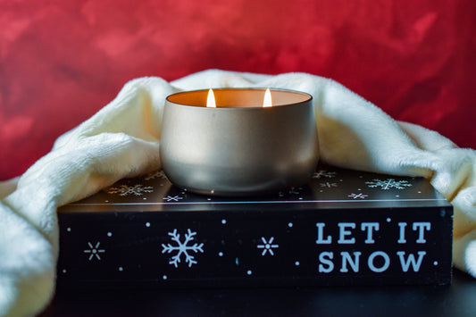 Christmas in July candle. Lit Gold bubble candle tin sitting on a prop that says Let it Snow. Red bakcground