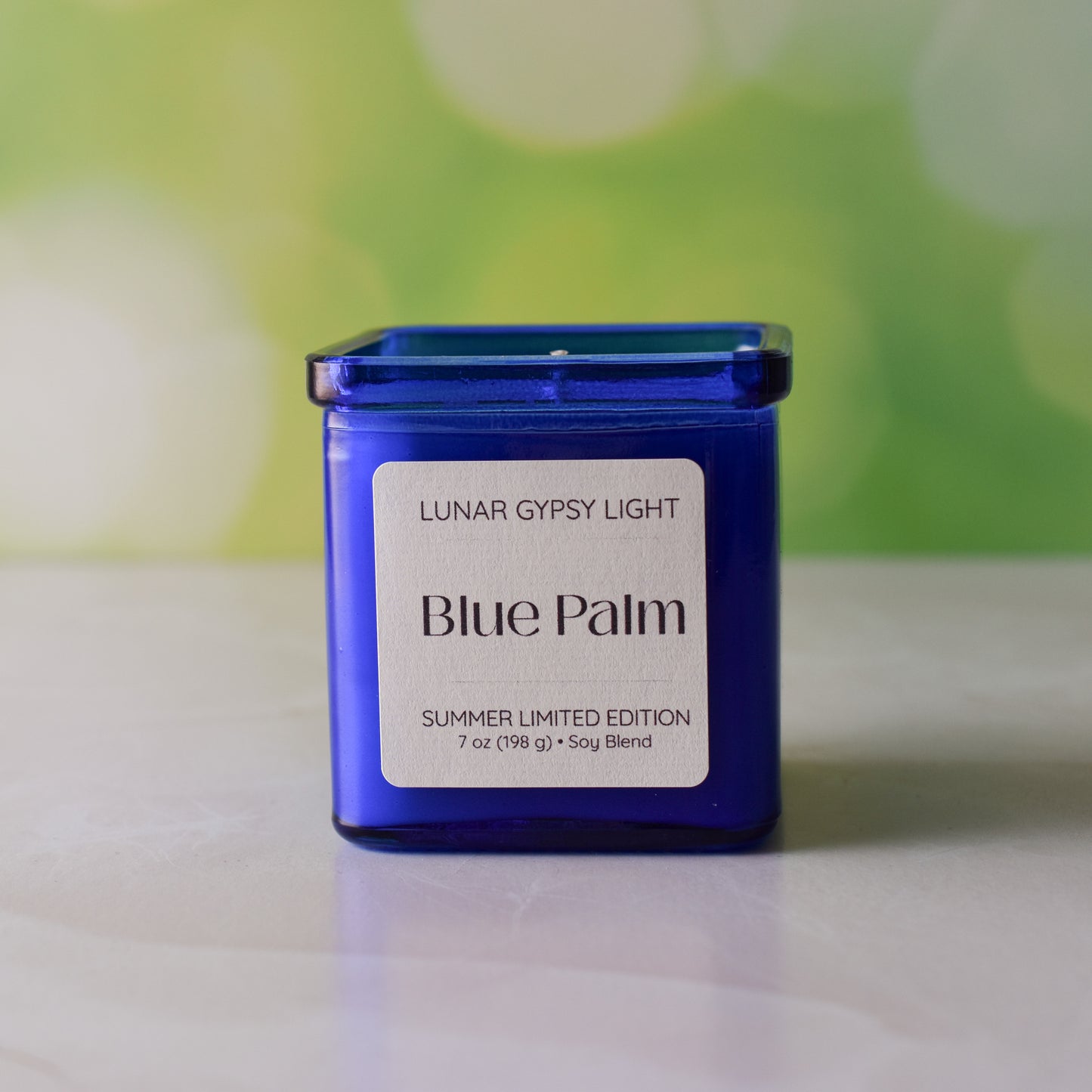 Blue candle labeled 'Blue Palm' by Lunar Gypsy Light on a white surface with a blurred green background