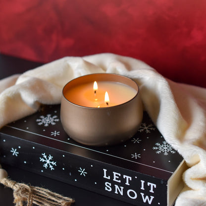 Lit candle with 2 flames in a gold bubble tin sitting on a book labled Let it Snow with a red background