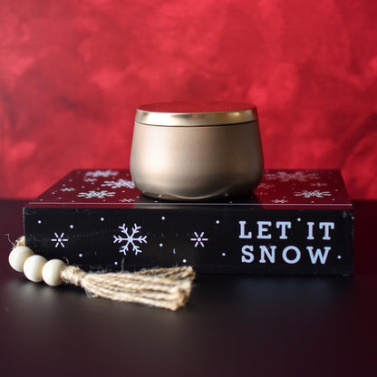 Candle in gold bubble tin with red background sitting on a book labeled let it snow