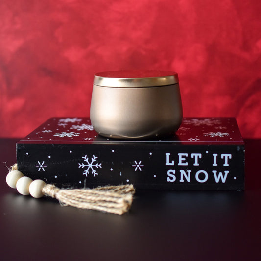 Candle in gold bubble tin with red background sitting on a book labeled let it snow