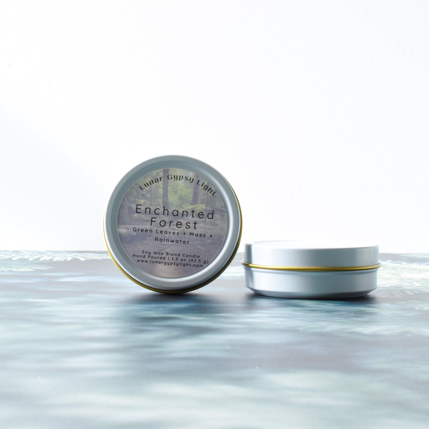 Two tins labeled 'Enchanted Forest' mini candle on a fir inspired surface with a white background