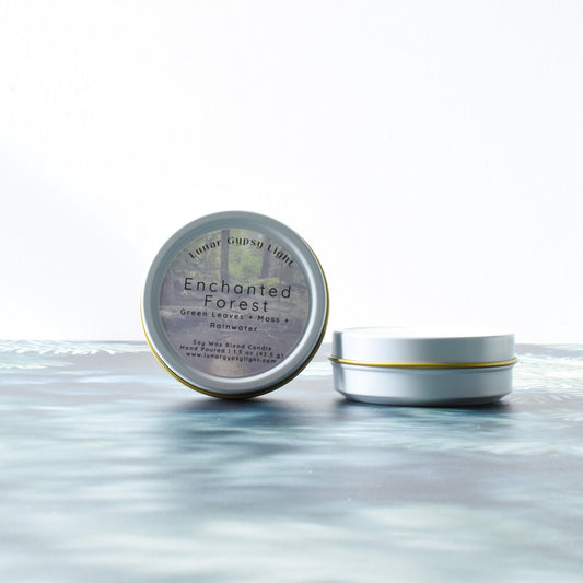 Two tins labeled 'Enchanted Forest' mini candle on a fir inspired surface with a white background