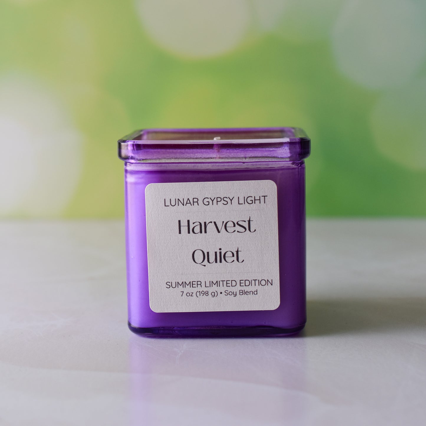 Purple candle labeled 'Harvest Quiet' by Lunar Gypsy Light on a white surface with a blurred green background